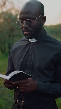 Medium vertical footage of African American priest in glasses performing burial service reading prayer book and holding beads at outdoor funeral ceremony
