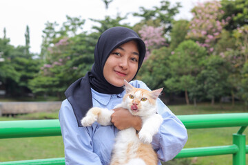 Outdoor portrait of Asian hijab woman holding and giving gentle touch to cat, taking care of her pet in nature park. Love relationship between humans and animals.