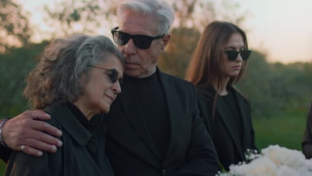 Side medium footage of Caucasian senior woman holding back tears mourning deceased and man consoling her standing by wooden coffin at outdoor funeral ceremony
