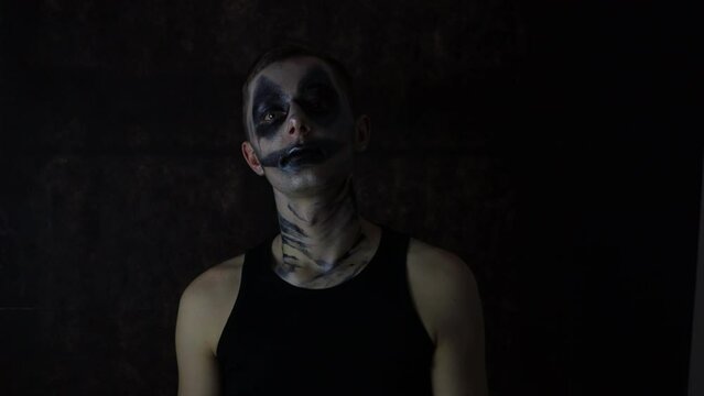 Young man with a scary Halloween dark clown makeup standing in front of a black background