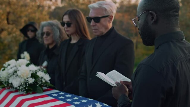 Rear footage of Black priest giving funeral rites reading prayer book to pay tribute to American soldier and Caucasian family standing by wooden coffin at cemetery