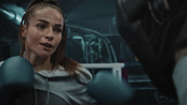 Close up of female fighter in boxing gloves practicing fighting technique and exercising in gym before fighting championship. Boxer hits punch mitts on boxing ring. Physical activity and workout.