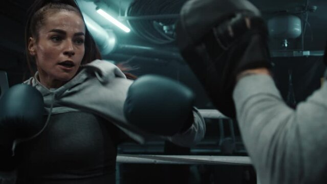 Close up of athletic woman hitting punching mitts while training in boxing gym. Female boxer in boxing gloves exercises with male trainer before fighting competition. Physical activity and workout.