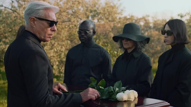 Side tracking footage of Caucasian senior man coming up to wooden casket and putting white rose blossoms on lid during outdoor funeral ceremony