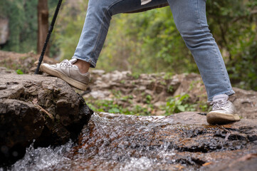 A female traveler with a trekking pole crossing water in a small canal while hiking on the mountain.