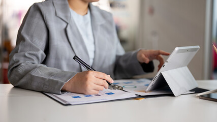 A businesswoman is working at her desk in the office, using her digital tablet and examining report.