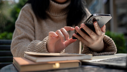 Cropped shot of an Asian woman using her smartphone while working remotely at a table outdoors.