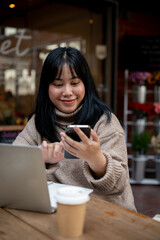 Positive Asian woman is responding to messages on her phone while working remotely at a coffee shop.