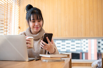 A positive Asian woman is sipping coffee and reading texts on her phone at a coffee shop.