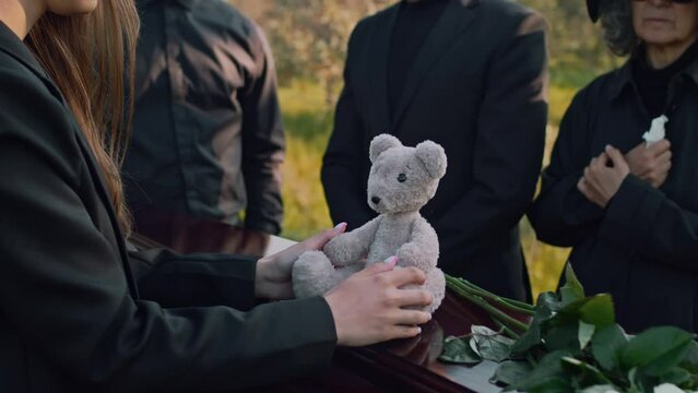 Side midsection footage of unrecognizable woman putting teddy bear on coffin lid during outdoor funeral for child
