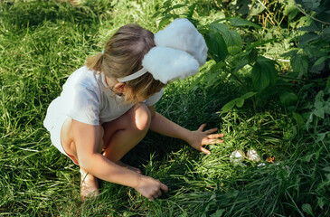 Child girl in a headband with bunny ears is looking for Easter eggs in the grass. Happy Easter religious holiday concept