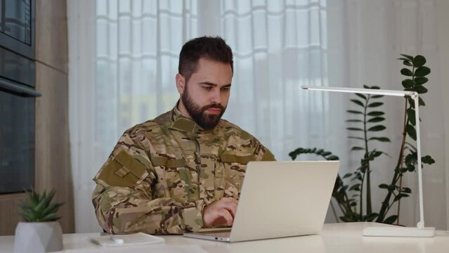 Male army soldier in military uniform using portable computer for emailing and working while sitting at home. Focused caucasian man typing at laptop keyboard and sending messages to commander office.