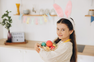 A cute little girl wearing plush bunny ears and apron holding basket full of Easter colorful eggs