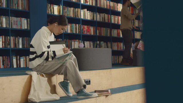 Full shot of Asian female student in striped jumper, pants and plimsolls sitting on bench in library with textbooks and writing in notebook, and group of young people chatting in background
