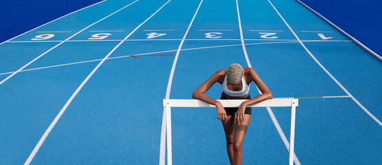 An African American female athlete is training on the Olympic track, leaning against the hurdles,...