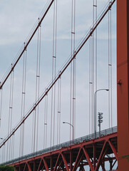 A close-up view of a part of a red suspension bridge against the backdrop of a cloudy sky.