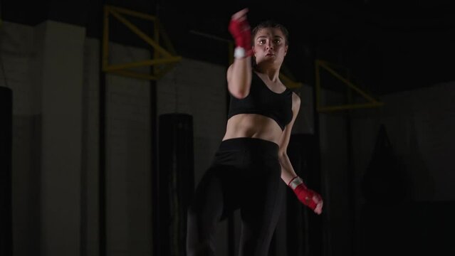 Woman fighter trains his kicks, training in the boxing gym, young woman looks at the camera and trains a series of punches, 4k slow motion.