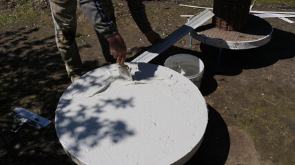 covering a round tabletop with plaster in the process of making original furniture using mortar and spatulas, the master spreads the mixture with a trowel onto the surface of the product outdoors
