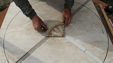 The tiler's hands place a square piece of material with a floral pattern on the smooth surface of a large light-colored tile, assembling a mosaic when tiling a table surface