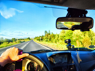 Car salon, steering wheel, hand of woman and view on nature landscape. View from seat of driver on Road, forest, blue sky, white clouds at sunny day. Concept of single trip of female traveller