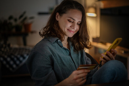 Young woman using smartphone and bank card shopping online at home in evening