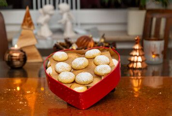 On a Christmas decorated table is a heart-shaped cookie box with round vanilla cookies dusted with powdered sugar. In the background two plaster angels are kissing.
