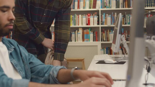 Medium close-up shot of busy Arab male student sitting at desktop computer in college library, thinking and typing on keyboard, unrecognizable man entering, taking out notebook and preparing to work