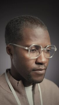 Vertical chest up slowmo portrait of young African American man wearing eyeglasses and looking at camera standing on grey background in studio