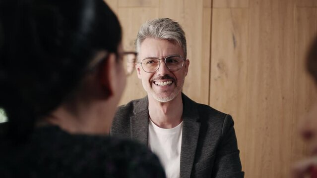 Handsome businessman talking to colleagues. gray-haired businessman wearing eyeglasses sitting in conference room and having friendly conversation with female colleagues sitting opposite