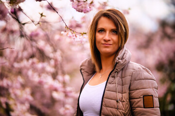 A young woman stands in a sea of ​​cherry blossoms and smiles into the camera. Cherry blossoms on the tree in full pink bloom.