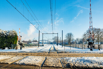 Snow covered train tracks at railway crossing, raised protective barriers, electric cables, bridge...