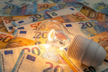 Light bulb lit, above euro banknotes. Electricity cost and tariffs.