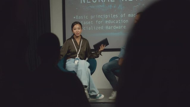 Full selective focus shot of Caucasian male speaker and Asian female moderator sitting together on stage at IT conference, finishing presentation, and audience clapping