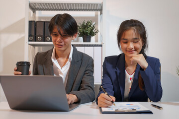 An Asian male consultant and an African American female intern sit at a table with laptops doing paperwork together to discuss a project's financial reports. Company business collaboration concept
