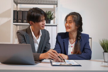 Fototapeta premium An Asian male consultant and an African American female intern sit at a table with laptops doing paperwork together to discuss a project's financial reports. Company business collaboration concept