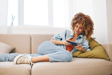 Happy woman sitting on a cozy sofa at home, holding her smartphone and smiling while enjoying online games and reading messages in a relaxed and cheerful atmosphere