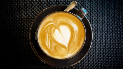 Delicious golden flat white Timorese coffee with love heart shape in modern black cup and saucer, top down birds eye view