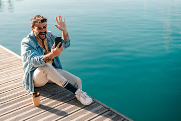 A trendy guy is sitting on a dock near sea and waving at the phone during his video call on the...