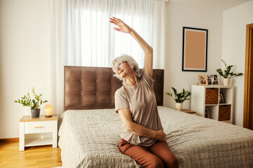 A senior woman is sitting on a bed in bedroom and doing morning gymnastic. - obrazy, fototapety, plakaty