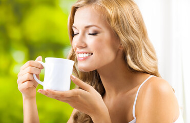 Portrait image - happy smiling woman holding white cup near window, at home house. Cheerful amazed girl with closed eyes, smelling aroma coffee drink indoor. Morning relax calm concept.