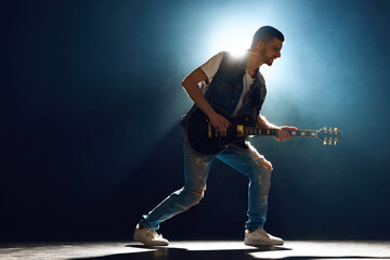 Full length portrait of guitarist in denim playing electric guitar, with dynamic backlight and...