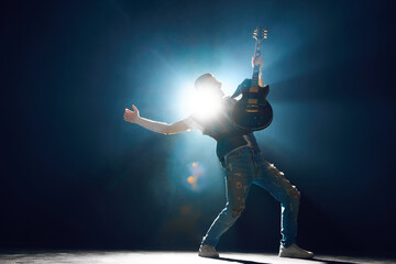 Rock performer in deep stance playing electric guitar on stage with backlit and smoke which...