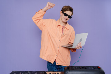 Cool young boy DJ dancing with laptop, in stylish clothes, glasses on a color background.