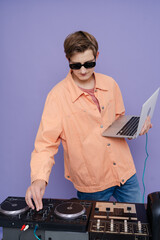 Teenager  DJ mixes music on a mixing console and laptop, in stylish clothes and glasses on a color background.