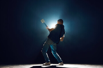 Performer with electric guitar leaning into musical moment against dark background with spotlights behind him. Concept of Rock-n-roll, music and dance, festivals and concerts, culture. - obrazy, fototapety, plakaty