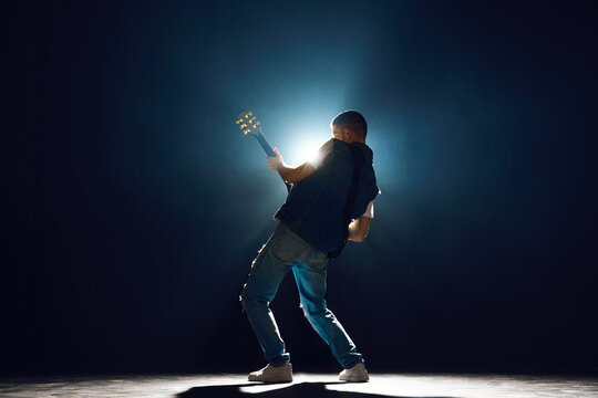 Performer with electric guitar leaning into musical moment against dark background with spotlights behind him. Concept of Rock-n-roll, music and dance, festivals and concerts, culture.