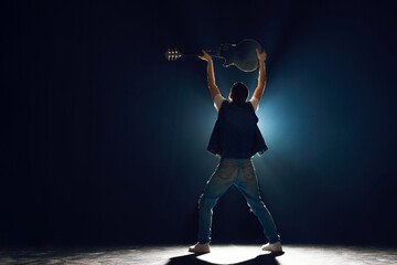 Rear view portrait of man in denim outfit holds electric guitar under head stands on stage....