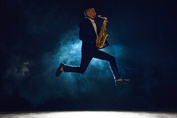 Dynamic shot of artistic man, solo performer jumping while playing jazz melody on saxophone against...