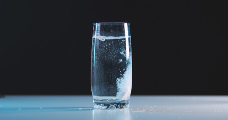 Effervescent tablet painkiller falls into glass of water on table against black background,...