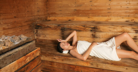 Young woman in towel relaxing in wooden sauna at spa. Asian woman in bathrobe doing body treatment...